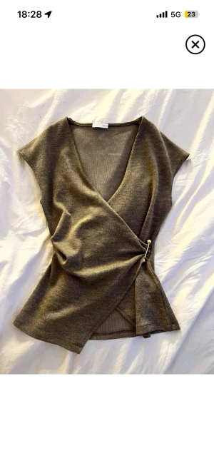 Brun omlottopp från Zara - Säljer en brun omlottopp från Zara i stickat material. Toppen har v-ringning, är ärmlös och stängs med en snygg guldfärgad säkerhetsnål på sidan. Perfekt för dig som gillar stilrena och trendiga plagg med en twist.