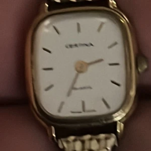 Certina damklocka i guld och silver - Snygg damklocka från Certina med rektangulärt guldtonat boett och vit urtavla. Armbandet är i rostfritt stål med guldfärgade detaljer och har ett coolt mönster. Quartz-urverk och minimalistiska indexmarkeringar. Perfekt accessoar för dig som gillar klassisk stil.