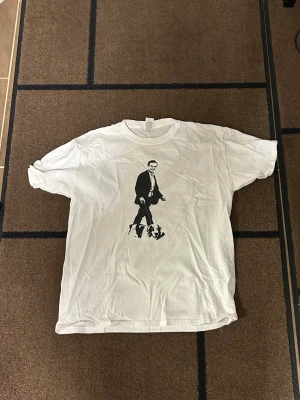 Ulf Kristersson T-shirt - Säljer en vit t-shirt från Gildan med ett stilrent svart tryck på framsidan som föreställer en person i kostym. T-shirten har klassisk rund hals och korta ärmar. Materialet är mjuk bomull och passformen är normal. Perfekt för dig som gillar grafiska motiv. 