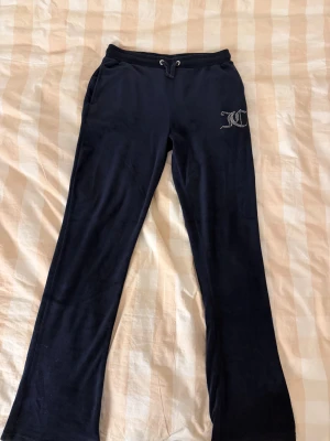 Mörkblå Juicy Couture byxor  - Snygga mörkblå mjukisbyxor från Juicy Couture med strassprydd JC-logga framtill. Byxorna har snörning i midjan, raka ben och två bakfickor. Mjukt och skönt material som ger en chill vibe, perfekt för att hänga med kompisar. Sömen har släppt på vissa ställen på bakfickorna, men läggs inte märke till.