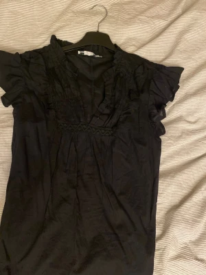 Svart blus med volang från lager 157 - Snygg svart blus från lager 157 med korta volangärmar och v-ringad hals. Blusen har dekorativa veck och spetsdetalj under bysten. Tillverkad i lätt och luftig bomull, perfekt för en trendig look. Passar dig som gillar romantiska detaljer och enkel stil. (Ser lite skrynklig ut men aldrig använt!)