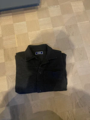 Mörkgrå overshirt från Jack & Jones - Säljer en stilren mörkgrå overshirt från Jack & Jones. Skjortjackan har klassisk krage, knappstängning framtill och en bröstficka. Materialet är mjukt och känns som bomull med en lätt melerad yta. Perfekt att slänga på över en t-shirt eller en tröcktröja för en chill och stilren look.
