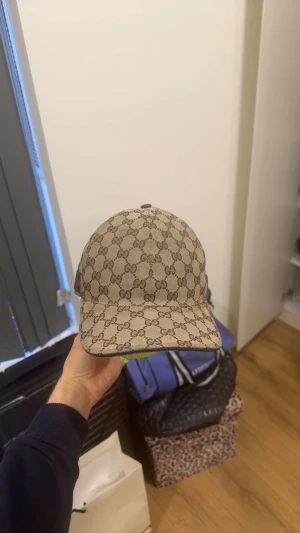 Gucci beige keps med GG-mönster Äkta - Snygg beige keps från Gucci med klassiskt GG-monogram över hela kepsen. Har grönt och rött band på sidan och justerbar rem baktill i mörkbrunt läder. Perfekt för dig som vill ha en lyxig och trendig accessoar med streetkänsla.