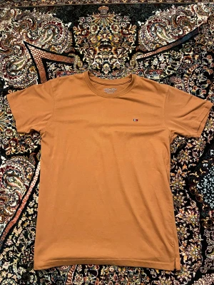 Brun t-shirt från Tommy Hilfiger - Snygg brun t-shirt från Tommy Hilfiger med klassisk rund hals och liten broderad logga på bröstet. T-shirten är i mjuk bomull och har en normal passform med korta ärmar. Perfekt för en clean och stilren look.