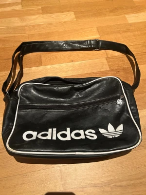 Svart axelväska från Adidas - Cool svart axelväska från Adidas med vit logga och vita detaljer. Väskan har ett stort fack och en praktisk dragkedja framtill. Perfekt för att bära med sig allt man behöver och har en retro vibe med klassisk Adidas-design.