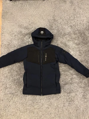 Mörkblå dunjacka från Sail Racing - Säljer en mörkblå dunjacka från Sail Racing med svarta detaljer på bröst och huva. Jackan har huva, två bröstfickor med dragkedja och Sail Racing-logga på bröstet. Insidan är fodrad och jackan har en snygg, sportig look som passar perfekt när det är kallt ute. Den är i jätte bra skick 