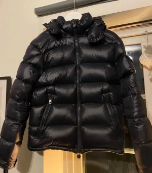 Svart Moncler pufferjacka med huva - Säljer en svart pufferjacka från Moncler med ikonisk logga på ärmen och detaljerad dragkedja. Jackan har huva, två sidofickor med dragkedja och en stilren, glansig finish. Perfekt för kalla dagar och riktigt snygg till streetwear-stilen.