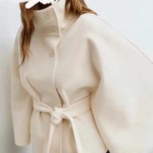  capejacka från Gina Tricot - Snygg capejacka i ljus beige/ vit från Gina Tricot med hög krage, knappar och knytbälte i midjan. Jackan har en stilren, vid siluett och är perfekt för dig som gillar minimalistisk design. Nyköpt och helt oanvänd. 