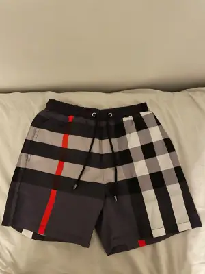 Snygga Burberry badshorts med klassiskt rutigt mönster i svart, vit, grå och röd. Shortsen har elastisk midja med svart snörning och metalländar med Burberry-logga. Bakficka och meshfoder på insidan. Perfekt för sommaren och strandhäng. Passade inte mig och är därför aldrig använd. Pris kan diskuteras 