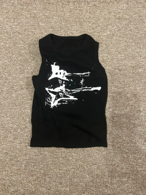 Svart croppad linnetopp med tryck - Svart croppad linnetopp med vitt grafiskt tryck framtill. Toppen är ärmlös och har en tight passform som sitter snyggt mot kroppen. Perfekt för dig som gillar en edgy och modern stil.