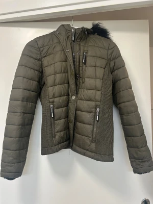 Superdry grön dunjacka - Mörkgrön dunjacka från Superdry med dubbla dragkedjor och knappa, storlek 40. Jackan har en avtagbar huva med fuskpäls. 