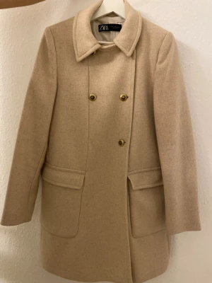 Beige kappa från Zara med guldknappar - Snygg beige kappa från Zara i storlek M. Klassisk modell med bred krage, dubbelknäppning med guldiga knappar och två stora fickor framtill. Tillverkad i ullblandning, perfekt för höst och vinter. Stilren och tidlös design som lyfter din look.