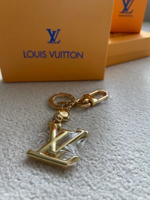 Vuitton  nyckelring i guld - Exklusiv nyckelring från  Vuitton i glänsande guldfärgad metall. Stor -logga som hänge, rund ring och karbinhake med ingraverad logotyp. Snygg och lyxig accessoar som sätter pricken över i:et på väskan eller nyckelknippan.
