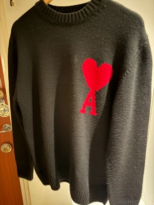 Svart stickad tröja från Ami Paris - Svart stickad tröja från Ami Paris med röd hjärtlogga och bokstaven A på bröstet. Tröjan har rund hals, ribbade muddar och lång ärm. Perfekt för dig som gillar stilrena plagg med en twist. Materialet är mjukt och håller värmen.