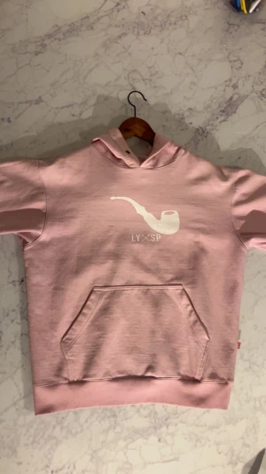 Rosa hoodie från Blaze S - Säljer en ljusrosa hoodie från Blaze i storlek S. Hoodien har en stor ficka framtill, huva med metallöljetter och ett vitt tryck av en pipa samt texten LY×SP på bröstet. Mjuk insida i fleece och klassisk passform. Perfekt för dig som gillar streetwear och snygga detaljer.