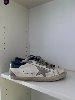 Golden Goose skor - Golden Goose sneakers i vitt skinn med grå stjärndetalj på sidan och mörkblå häl. Skorna har grå snörning och platt sula, samt GGDB/SSTAR-tryck i blått. OBS! ⚠️ Skorna har större defekter på sula (skriv privat för fler bilder). 