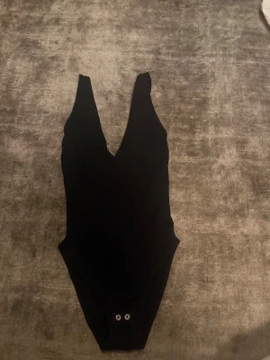 Svart ärmlös bodysuit med djup v-ringning - Säljer en svart bodysuit med djup v-ringning och breda axelband. Bodyn är ärmlös och har tryckknappar i grenen för enkel på- och avtagning. Materialet känns mjukt och stretchigt, perfekt för att styla med jeans eller kjol. Enkel och stilren design.