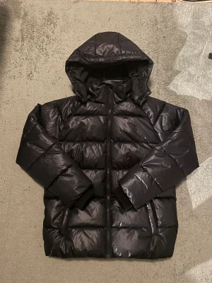 Svart pufferjacka från Kappahl - Svart pufferjacka från Kappahl med huva och dragkedja framtill. Jackan har quiltad design och två sidofickor. Materialet är syntet och polyester, vilket gör den lätt och varm. Perfekt för kalla dagar och har en stilren look som funkar till det mesta.