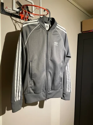 Grå Adidas sst med vita ränder - Säljer en grå Adidas sst med klassiska vita ränder längs ärmarna och Adidas-logga på bröstet. Jackan har dragkedja framtill, ribbade muddar och krage. Perfekt för dig som gillar sportig och stilren look.
