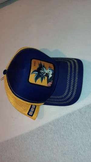 Capslab Dragon Ball Z Goku keps - Snygg blå och gul truckerkeps från Capslab med Goku-motiv från Dragon Ball Z framtill. Kepsen har mesh-bakstycke i gult, böjd skärm med dekorativa sömmar och justerbar snapback. Materialet är polyester och bomull, perfekt för animefans. Köpt från Tyskland för 120 Euro. Och säljs för att jag inte har använding av den.