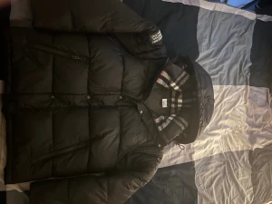 Svart Burberry pufferjacka med huva - Säljer en svart pufferjacka från Burberry med klassiskt rutigt foder i huvan. Jackan har dragkedja, tryckknappar och två sidofickor med dragkedja. Perfekt för kalla dagar med sin tjocka och quiltade design. Stilsäker look med Burberry-logga på ärmen. Har använts i mindre 1 månad.