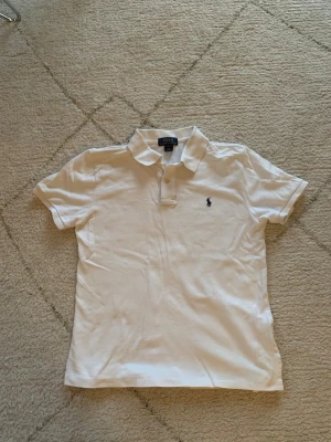 Vit pikétröja från Polo Ralph Lauren - Vit pikétröja från Polo Ralph Lauren med klassisk krage och två knappar framtill. Tröjan har det ikoniska broderade Polo-logot på bröstet och är tillverkad i mjuk bomull. Custom fit-modell som sitter snyggt och bekvämt. Perfekt för en clean och stilren look.