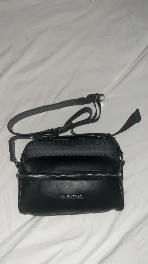 Svart axelväska från Mario Valentino - Snygg svart axelväska från Mario Valentino med justerbar rem och silverfärgad dragkedja. Väskan har logotypen VALENTINO framtill och diskret mönster med text på ovandelen. Tillverkad i skinnliknande material och levereras med original dustbag.