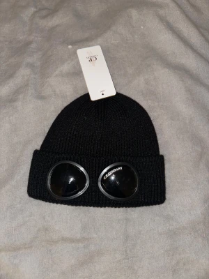 Svart och vit C.P. Company gogglemössa - Två stickade mössor från C.P. Company, en svart och en vit, med ikoniska goggle-linser på uppviket. Mössorna är ribbstickade och har rundad form. Linserna är svarta med C.P. Company-logga. Perfekt för dig som gillar streetwear och vill sticka ut.