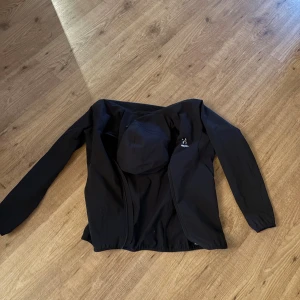Svart vindjacka från Haglöfs med luva  - Svart vindjacka från Haglöfs med stilren design och praktisk huva. Jackan har dragkedja framtill, elastiska ärmslut och diskret logga på bröstet. Perfekt för blåsiga dagar och outdoor-aktiviteter. Materialet är lätt och syntetiskt för smidig rörelse.