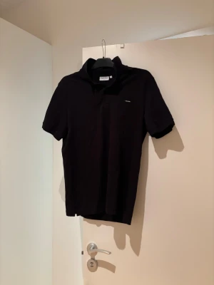 Svart pikétröja Calvin Klein Slim Fit - Svart pikétröja från Calvin Klein i slim fit-modell. Klassisk krage och korta ärmar, med diskret logga på bröstet. Tillverkad i mjuk bomull som är skön mot huden. Perfekt för dig som gillar stilrena och enkla plagg.