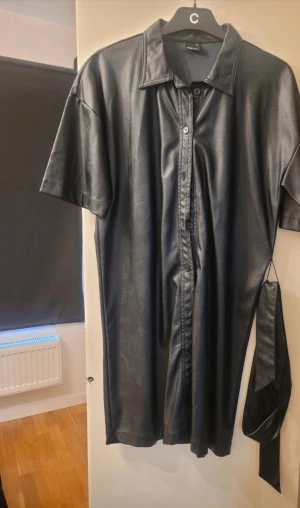 Svart skjortklänning i skinn från Gina Tricot - Cool svart skjortklänning från Gina Tricot i storlek 36. Klänningen har korta ärmar, klassisk krage och knappar hela vägen framtill. Tillverkad i mjukt syntetskinn och med tillhörande bälte för att markera midjan. 