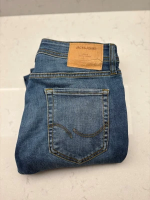 Blå jeans från Jack & Jones - Snygga blå jeans från Jack & Jones med klassisk femficksdesign och kontrastsömmar. Jeansen har en läderpatch med logga baktill i midjan och är tillverkade i slitstarkt denimtyg. Perfekta för dig som gillar en enkel och tidlös look.