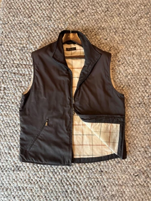 Loro piana väst - Brun väst från Loro Piana med hög krage och två fickor med dragkedja. Insidan har ett stilrent rutigt mönster i beige, grått och rött. Materialet är polyester och polyuretan, vilket gör den lätt och bekväm. Perfekt för lager-på-lager och snygg till jeans eller chinos.