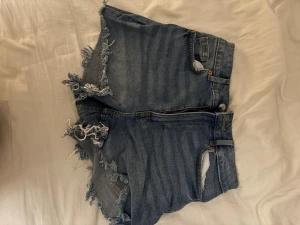 Blå jeansshorts med fransar från H&M Divided - Säljer ett par blå jeansshorts från H&M Divided i storlek 34. Shortsen har råa, fransiga kanter och klassisk femficksdesign med dragkedja och knapp framtill. Perfekt för dig som gillar en avslappnad och edgy stil.