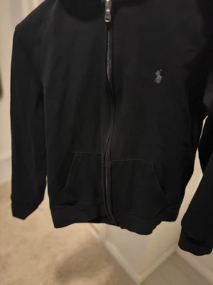 Svart hoodie från Polo Ralph Lauren - Svart hoodie med dragkedja från Polo Ralph Lauren. Klassisk design med två fickor fram, broderad logga på bröstet och huva. Tillverkad i mjuk bomullsblandning som är skön att ha på sig. Perfekt för en chill och stilren look. Det ända felet med tröjan är att en liten del av dragkedjan har gått av men det är inget större problem.
