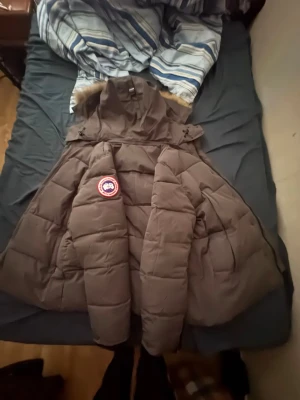 Mörkgrå Canada Goose dunjacka med päls - Säljer en mörkgrå dunjacka från Canada Goose med fet päls på huvan och klassisk logga på ärmen. Jackan är quiltad, har lång ärm och dragkedja framtill. Insidan är fodrad med blankt syntetmaterial. Perfekt för kalla vinterdagar.