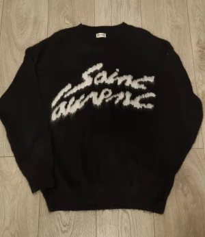 Svart Saint Laurent stickad tröja - Svart stickad tröja från Saint Laurent med vit, suddig logotyp på bröstet. Tröjan har rund hals, ribbade muddar och lång ärm. Materialet är mjukt och fluffigt, troligen mohair eller ullblandning. Perfekt för dig som gillar exklusiva streetwear-vibbar.