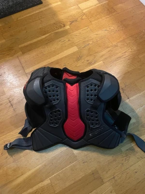 Svart och röd hockeyaxelskydd CCM - Säljer ett fett hockeyaxelskydd från CCM i svart och rött med coola detaljer och robust design. Skyddet har justerbara remmar och extra vaddering för axlar och bröst. Perfekt för dig som vill ha max skydd på isen och snygg stil.