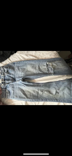 Blå jeans från Gina med slitningar - Säljer ett par ljusblå jeans från Gina tricot i storlek 36. Byxorna har en klassisk femficksdesign och coola slitningar för en trendig look. 