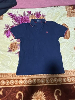 Marinblå Fred Perry pikétröja M - Snygg marinblå pikétröja från Fred Perry i slim fit. Tröjan har klassisk krage med röda och vita ränder, korta ärmar med matchande kant och den ikoniska Fred Perry-loggan broderad på bröstet. Perfekt för dig som gillar stilren och sportig look.