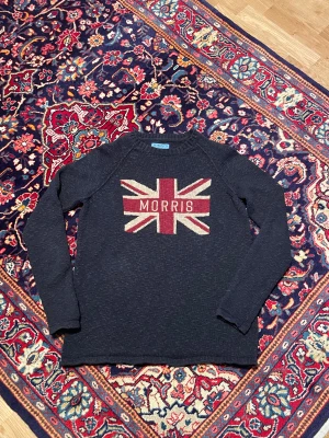 Mörkblå stickad tröja Morris Co - Snygg mörkblå stickad tröja från Morris Co i storlek medium. Framsidan har ett Union Jack-inspirerat mönster i rött, beige och mörkblått med texten 'Morris' i mitten. Tröjan har långa ärmar och rund hals, perfekt för dig som gillar klassisk stil med en twist.
