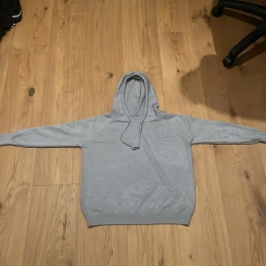 Ljusgrå hoodie med huva och snörning - En stilren ljusgrå hoodie med huva och snörning. Tröjan har långa ärmar och ribbade muddar vid ärmslut och nederkant. Perfekt för en chill och avslappnad look. Materialet känns mjukt och bekvämt, troligtvis bomull eller en bomullsblandning.