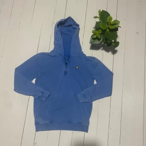 Blå 1/4 zip hoodie från Lyle & Scott - Snygg blå hoodie från Lyle & Scott med halv dragkedja och klassisk gul fågel-logga på bröstet. Mjuk bomullskänsla, ribbade muddar och stor huva. Perfekt för chill dagar och streetwear-stil. Axel bredden är 50cm. Längden är 62cm. Ärmarna är 58cm. 