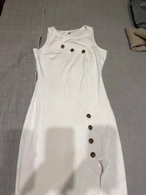 Vit ärmlös klänning med knappar - Snygg vit klänning. Dekorerad med guldiga knappar både upptill och längs sidan. Använd endast en gång.