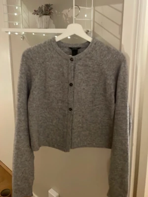 Grå kofta från H&M med knappar - Mysig grå kofta från Lindex med rund hals och tre svarta knappar framtill. Koftan är långärmad och har en mjuk, stickad känsla som passar perfekt för kyliga dagar. Enkel och stilren design som funkar till många olika outfits.