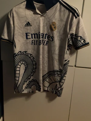 Real Madrid x Adidas draktröja - Säljer en Real Madrid tröja från Adidas med coolt drakmönster i grått och marinblått. Tröjan har korta ärmar, krage och detaljer i guld. Tryck med klubbmärke och sponsor framtill. Perfekt för dig som gillar fotboll och vill sticka ut på läktaren.