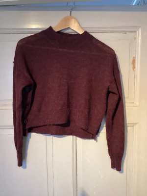 Vinröd stickad tröja med knappar - Säljer en vinröd stickad tröja med lång ärm och hög hals. Tröjan har en croppad passform och snygga knappar längs ena sidan. Perfekt för dig som gillar en enkel men stilren look med lite extra detalj.