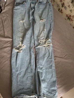 Levi's 501 ljusblå ripped jeans - Ljusblå Levi's 501 jeans med klassisk femficksdesign och slitna detaljer på knän och bakficka. Jeansen har rak passform och är tillverkade i robust bomullsdenim. Perfekt för dig som gillar en avslappnad och edgy stil.