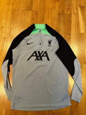 Nike Liverpool Dri-Fit träningströja M - Nike Liverpool FC långärmad träningströja i slim fit med Dri-Fit-teknologi. Tröjan är ljusgrå med svarta ärmar och detaljer, samt en mintgrön krage. Stor AXA-logga och LFC-märke på bröstet. Perfekt för fotbollsträning och sport.