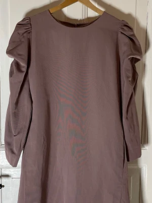 Ljuslila COS klänning med puffärm - Säljer en stilren dämpad rosa/lila klänning från COS i storlek 44. Klänningen har puffade långärmar och rak passform med rund halsringning. Materialet är lite tyngre och har dolda fickor i sidan.. Dragkedja bak för enkel påklädning. Perfekt för dig som gillar minimalistisk design.